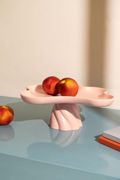 Three peaches on a pink, sculptural pedestal dish, with another peach and books on a glossy blue tabletop.