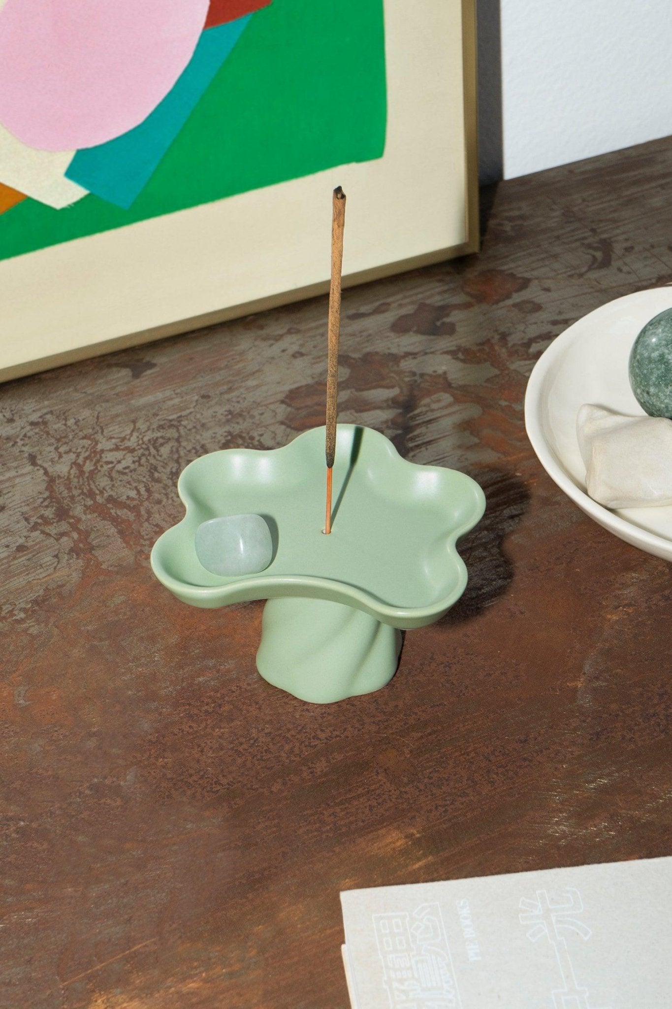 The Octaevo Templo Incense Holder, a green stoneware catchall, sits by art and a bowl on a metal table.