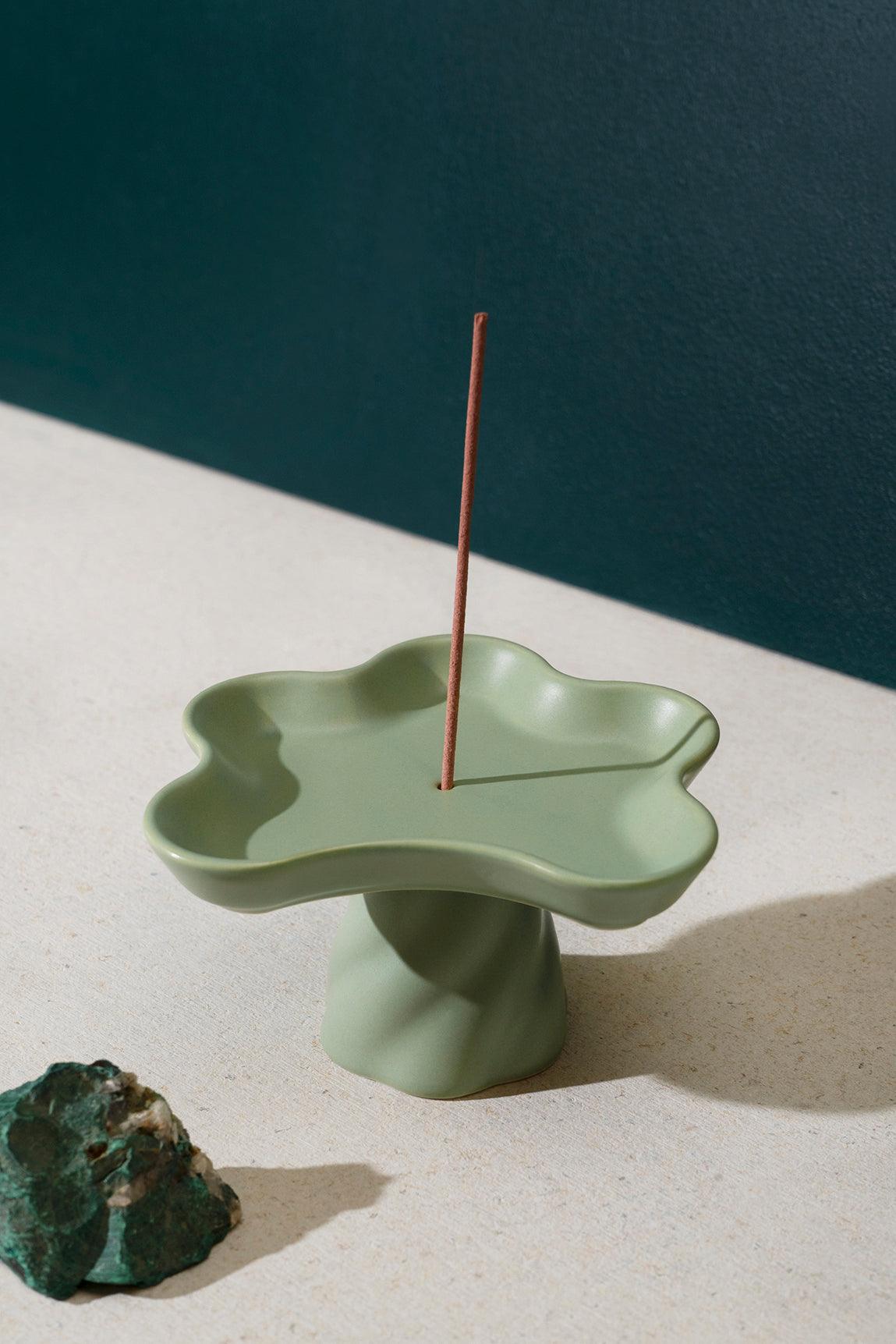 The Octaevo Templo Incense Holder, a green flower-shaped stoneware piece, holds a lit stick on a light surface.