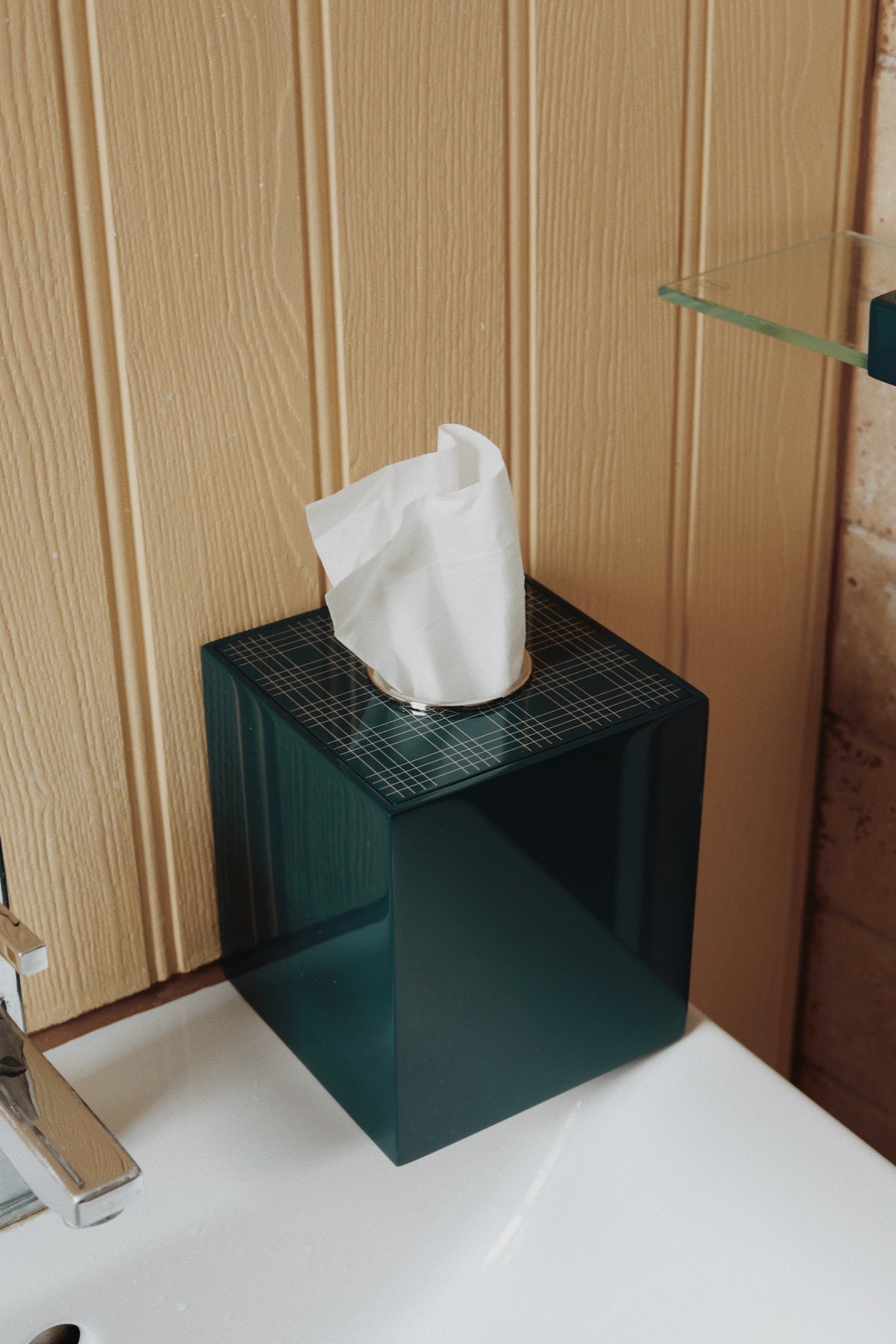 A dark teal translucent lacquered tissue box cover with a white tissue paper inserted, sitting on a white bathroom counter against a light wood paneled wall.