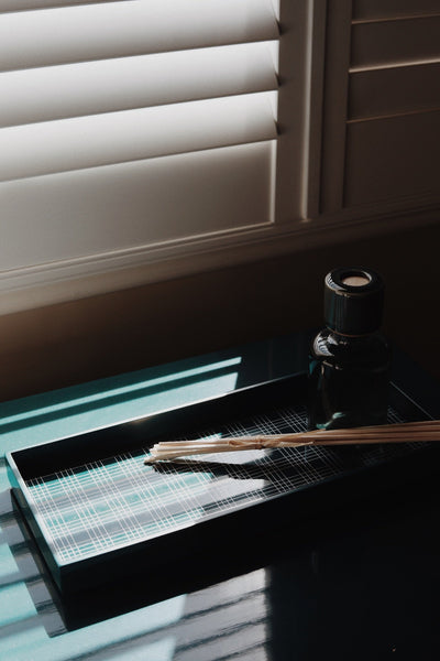 Vinidec slim linear lacquered tray in deep teal with white grid insert holding a glass reed diffuser in dramatic window light