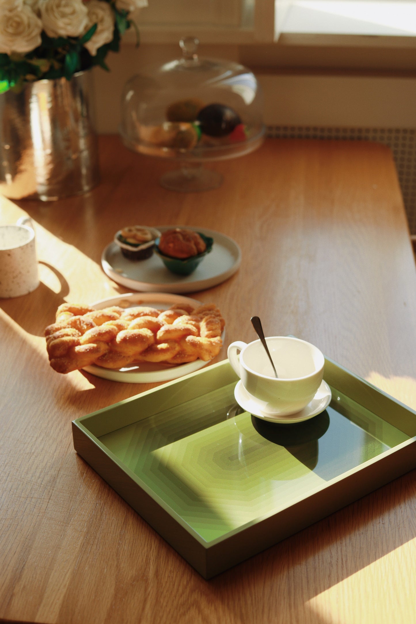 Medium green Chiaroscuro high-gloss lacquer tray holding a ceramic coffee cup and saucer on a wooden breakfast table with pastries in warm morning light