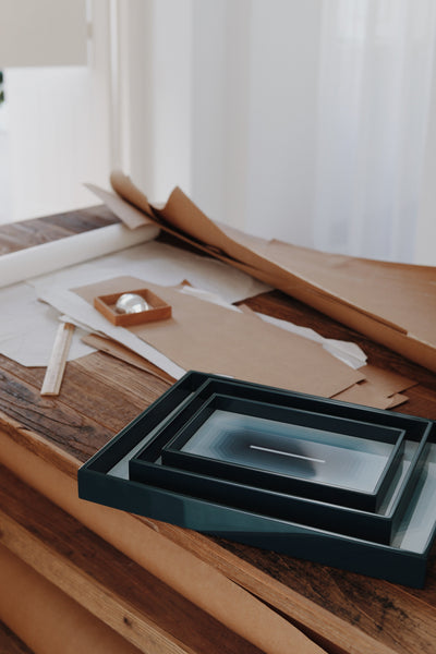Small blue Chiaroscuro high-gloss lacquer tray nested inside medium and large trays on a wooden studio table. Table styling
