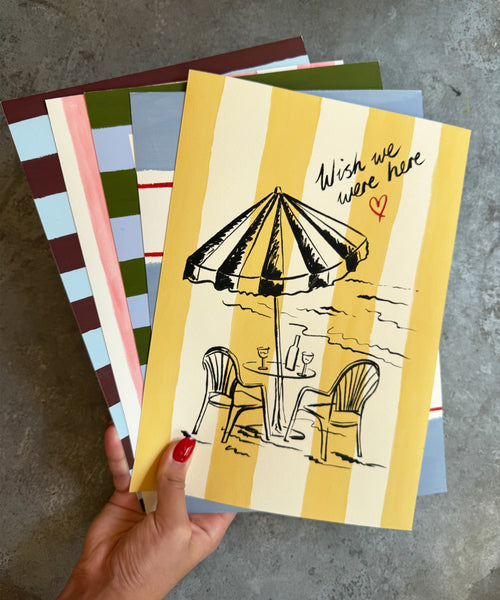 "Wish We Were Here" ink sketch print of a beach parasol and plastic chairs on a striped background, with the rest of seaside