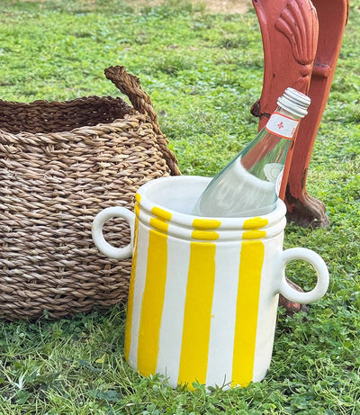 A ceramic wine cooler with white and yellow stripes, featuring two handles, partially filled with a wine bottle, placed on grass next to a wicker basket.
