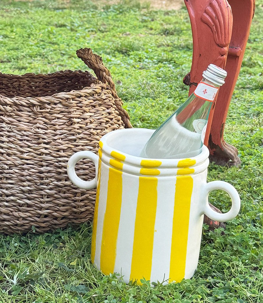 A ceramic wine cooler with white and yellow stripes, featuring two handles, partially filled with a wine bottle, placed on grass next to a wicker basket.